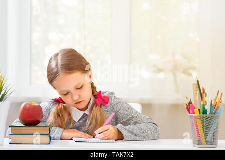 Una bambina studente si siede al tavolo e scrive in un notebook su uno sfondo sfocato. Il concetto di istruzione e la scuola. Foto Stock