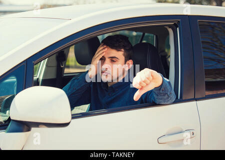 Esaurito giovane uomo seduto dietro al volante della vettura di esame che mostra il pollice verso il basso gesto mantiene la mano alla sensazione di testa mal di testa. Sfortunato s Foto Stock