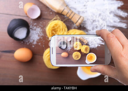 Le mani di uomo con lo smartphone tenendo photo uovo, farina, sale ingredienti per la pasta di spaghetti. Concetto di cucina a vista. Vista dall'alto. Foto Stock