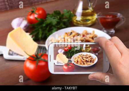 Le mani di uomo con lo smartphone tenendo foto penne piccante pasta bolognese con verdure, peperoncino e formaggio in salsa di pomodoro sullo sfondo di pomodori Foto Stock