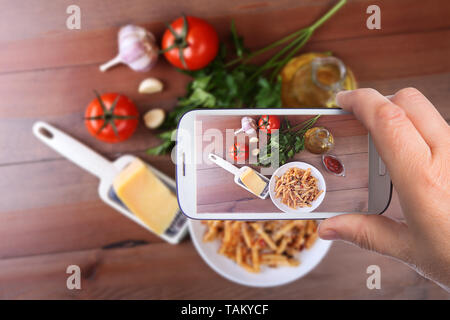 Le mani di uomo con lo smartphone tenendo foto penne piccante pasta bolognese con verdure, peperoncino e formaggio in salsa di pomodoro sullo sfondo di pomodori Foto Stock