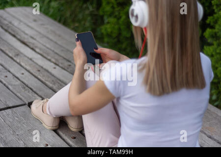 Torna ragazza con smartphone su gambe e cuffie seduta su una panchina in un parco e ascolto di musica. Una donna che indossa pantaloni di colore rosa e beige e scarpe di vernice Foto Stock