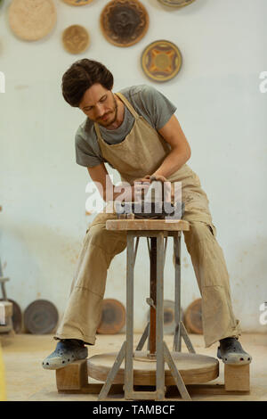 Elaborazione intensamente argilla. Focalizzato occupato uomo seduto su una tavola di legno ruota in ceramica e la produzione di nuova creazione Foto Stock