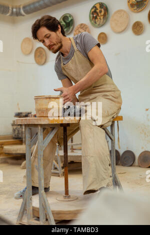 Trattare con argilla. Barbuto sperimentato uomo in grigio grembiule seduti sulla sedia alta e intensamente di argilla di elaborazione Foto Stock