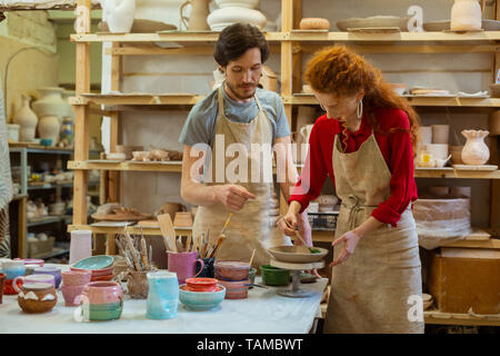 Aggiunta di pattern. Talentuoso dai lunghi capelli donna in grembiule con lo strumento pennello per decorare appena fatta la piastra Foto Stock