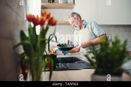 Un anziano uomo in ambienti interni in cucina a casa, lavaggio fino a pan. Foto Stock