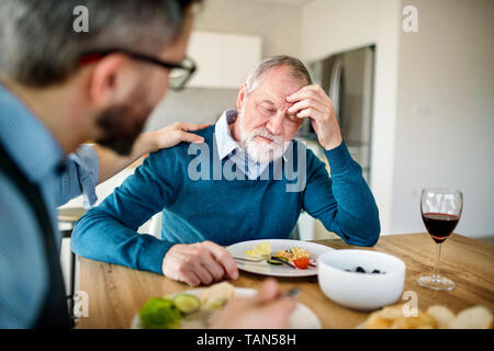 Un adulto hipster figlio e frustrato padre senior al chiuso in casa, mangiare pranzo leggero. Foto Stock