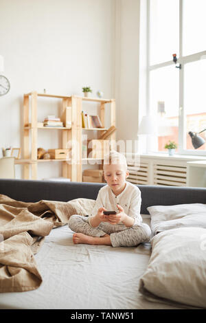 Gadget gravi-addicted kid seduta con le gambe incrociate sul divano letto e utilizzando dispositivo moderno mentre navigano in rete a casa Foto Stock