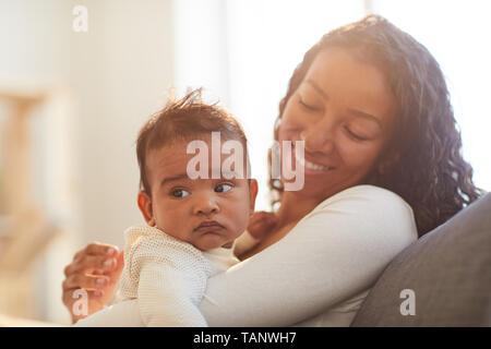 Felice parentesi eccitato dai capelli giovane donna africana seduti sul divano e trattenere il bambino in armi a casa Foto Stock