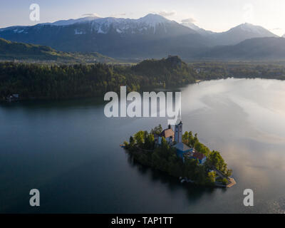 Sunrise vista aerea di St Marys chiesa sull isola del lago di Bled, Slovenia. Sullo sfondo vi è il castello di Bled su rocce e montagne distanti all'inizio su Foto Stock