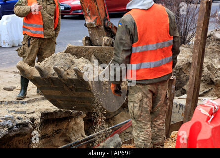 Una benna di una costruzione pesante escavatore circondato da operai stradali ascensori sabbia durante una fognatura riparazione su una strada della citta'. Foto Stock