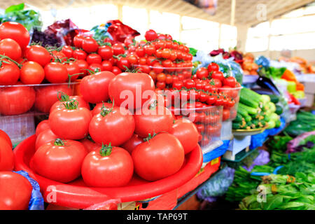 Diverse varietà di ripe pomodori rossi in background in una sfocatura di cetrioli, spinaci e altri ortaggi venduti nei vassoi sul mercato Foto Stock