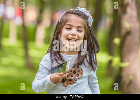 Adorabili e quattro anni carino bambina abiti casual detiene il cellulare mentre rideva di outdoor in posizione di parcheggio Foto Stock