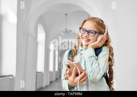 Bella, bella, positivo schoolgirl ascoltando musica con le cuffie durante la pausa scolastica. Bambino felice in piedi nel corridoio della scuola primaria. Foto Stock