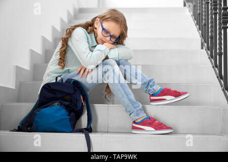 Piuttosto, triste schoolgirl in bicchieri seduto sulla scalinata di scuola, appoggiato sulle ginocchia. Premuto ragazza con lunghi capelli ricci guardando giù, pensando. Foto Stock