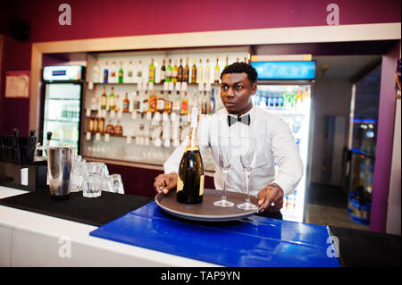 African American barista presso il bar tenendo con una bottiglia di champagne e bicchieri sul vassoio. Foto Stock