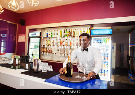 African American barista presso il bar tenendo con una bottiglia di champagne e bicchieri sul vassoio. Foto Stock