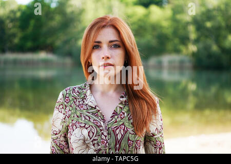 Raffreddare e fiducioso giovane donna indossa motivo floreale abiti estivi con lunghi capelli rossi in piedi dal lago - autentico persone reali
