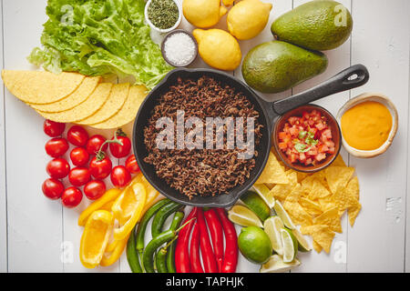 Chili con carne in padella su bianco tavolo in legno. Gli ingredienti per la realizzazione di Chili con carne.Vista dall'alto. Chili con carne, nachos, tacos, tigli, avocado, Foto Stock