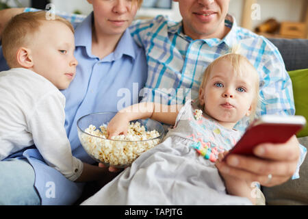 Close-up della famiglia giovane seduto sul divano e guardare la tv insieme mentre mangiare popcorn, focus su blue-eyed girl concentrata su cartoon Foto Stock