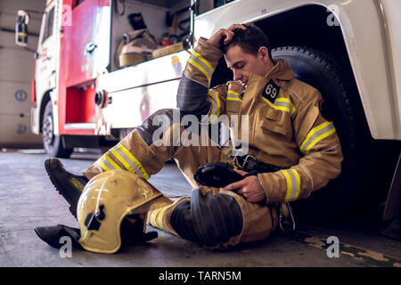 Foto di stanchi fireman seduto sul pavimento vicino al camion dei pompieri in corrispondenza della stazione Foto Stock