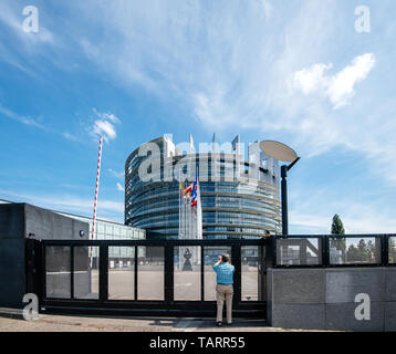 Strasburgo, Francia - 26 Maggio 2019: Seniopr uomo prendendo fotografia attraverso il cancello chiuso del Parlamento europeo sede con tutti Unione europea bandiere sventolano - cielo blu chiaro in background Foto Stock