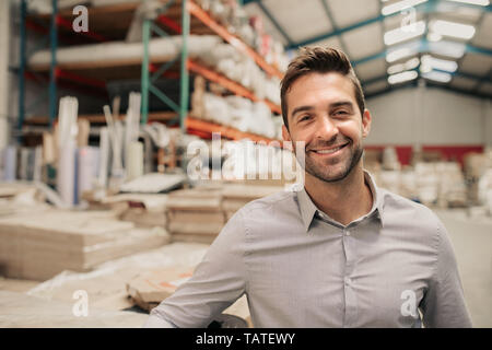 Ritratto di un sorridente warehouse manager appoggiata contro alcuni stock con pile di tappeti sui ripiani in background Foto Stock