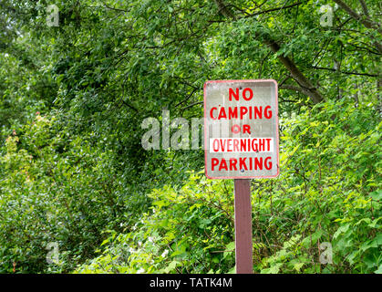 Nessun campeggio o parcheggio notturno segno di avvertimento sul montante con caratteri rossi sulla strada rurale accanto a verdi alberi. Foto Stock