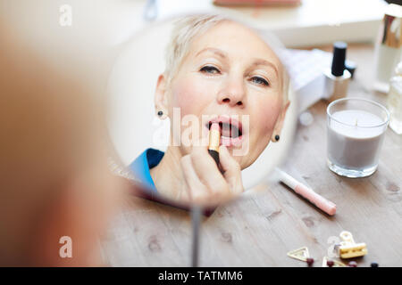 Di mezza età femmina bionda cercando in specchio mentre la gestione con il trucco e applicazione di rosa pastello rossetto Foto Stock