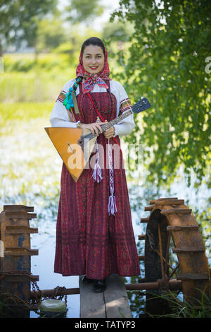 Giovane donna sorridente in tradizionali abiti russo sorge su un piccolo ponte in prossimità del lago, e tenendo la balalaika, vista verticale Foto Stock