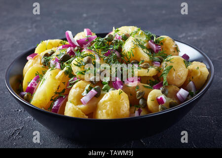 Tedesco saporiti insalata di patate con cipolla rossa, capperi, verdi e mostarda condimento di aceto in una ciotola di nero su un tavolo in cemento, vista da sopra Foto Stock