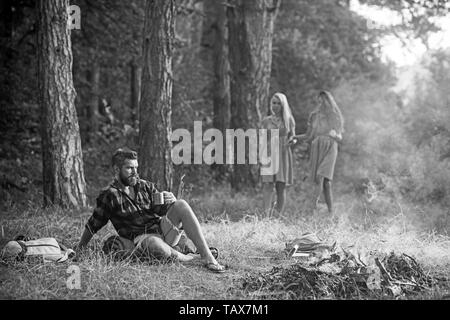 Il concetto di ambiente. Riflessivo uomo barbuto guardando le fiamme di fuoco. Due ragazze a piedi nudi ad esplorare la foresta. Foto Stock