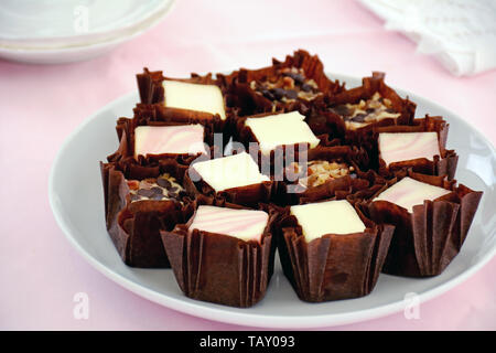 La vaniglia, lampone swirl e noce chocolate chip mini cheesecake morsi in involucri scuro sul round piastra bianca sul tessuto rosa sfondo. Foto Stock