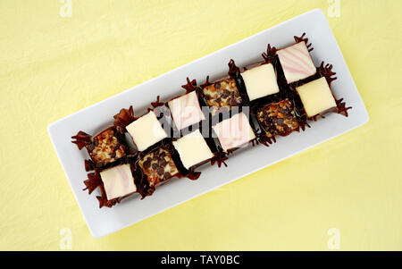 La vaniglia, lampone swirl e noce chocolate chip mini cheesecake morsi in involucri scuro sul lungo la piastra bianca sul tessuto giallo dello sfondo. Foto Stock