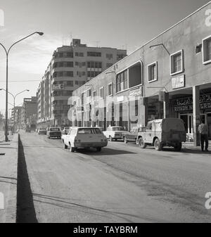 Anni sessanta, storico, diurno e una strada polverosa in Riyadh mostra edifici, vetture d'epoca e sulla destra alcuni negozi locali, Riad in Arabia Saudita. A partire da una popolazione di circa 150.000 in questa epoca la città capitale del paese è cresciuta convenientemente, con quasi 7 milioni di abitanti nel 2018, uno dei più grandi del mondo arabo. Foto Stock