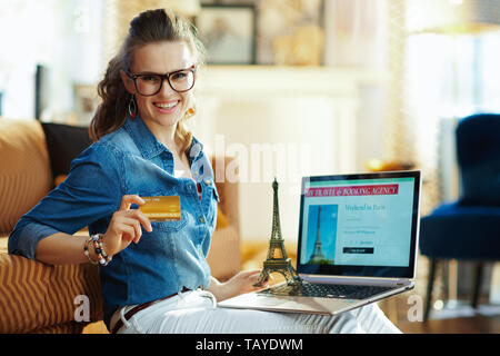 Felice giovane donna con aperto sul computer portatile online travel & BOOKING AGENCY sito e un souvenir della torre eiffel azienda carta di credito nella casa moderna Foto Stock