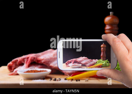 Le mani di uomo con lo smartphone tenendo foto Raw nervature su un rustico tagliere con sale e pepe e il macinino per spezie . Sfondo nero per la copia Foto Stock
