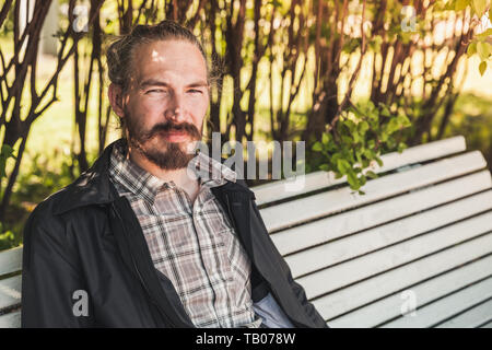Giovane uomo barbuto si siede su un banco di bianco in estate park, ritratto all'aperto Foto Stock