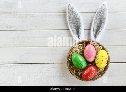 Colorate le uova di Pasqua e il coniglietto di pasqua orecchio coniglio in cestello nido decorazione bianco su sfondo di legno vista superiore Foto Stock