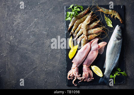 Materie tutta la spigola, gamberoni, calamari e le fette di limone e le spezie e le erbe su una piastra di ardesia su un tavolo in cemento, vista da sopra, close-up, flatlay, co Foto Stock