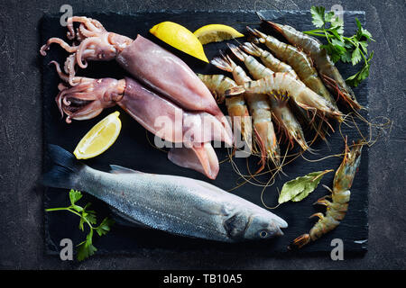 La spigola, gamberoni, calamari e le fette di limone e le spezie e le erbe su una piastra di ardesia su un tavolo in cemento, vista da sopra, close-up, flatlay Foto Stock
