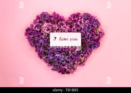 Forma di cuore fatta di viola fiori lilla con iscrizione ti amo sul rosa pastello sfondo. Simbolo d'amore. Copia dello spazio. Vista dall'alto. Biglietto di auguri Foto Stock