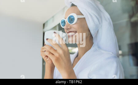 Bella donna in accappatoio indossando occhiali da sole e di bere il caffè. Sorridente giovane femmina avente il caffè dopo la doccia. Foto Stock