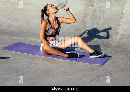 Donna seduta su un tappetino fitness e acqua potabile. donna in forma che beve l'acqua da una bottiglia dopo l'allenamento. Foto Stock