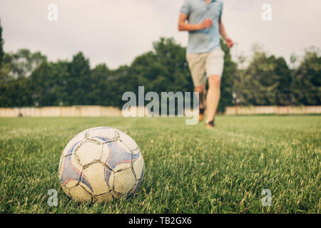 Pallone da calcio in erba su un campo sportivo con un anonimo giovane uomo in background in esecuzione verso di essa in un basso angolo di visione Foto Stock