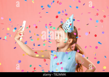 Allegro bambina festeggia il compleanno. Il bambino tiene il telefono, prende un selfie sotto la pioggia di coriandoli. Closeup ritratto su sfondo rosa. Foto Stock