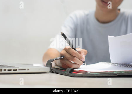 primo piano con la mano che fa il lavoro domestico con la penna e. computer portatile seduto sul tavolo Foto Stock