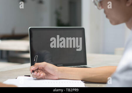 Vista posteriore studente facendo compiti a casa con il computer portatile e la scrittura in notebook presso il cafe, giovane uomo che lavora presso il cafe con notebook e laptop Foto Stock