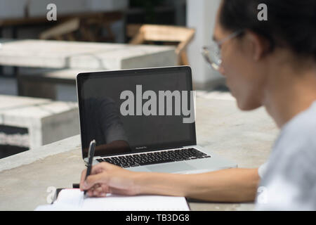 Vista posteriore studente facendo compiti a casa con il computer portatile e la scrittura in notebook presso il cafe, giovane uomo che lavora presso il cafe con notebook e laptop Foto Stock