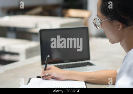 Vista posteriore studente facendo compiti a casa con il computer portatile e la scrittura in notebook presso il cafe, giovane uomo che lavora presso il cafe con notebook e laptop Foto Stock